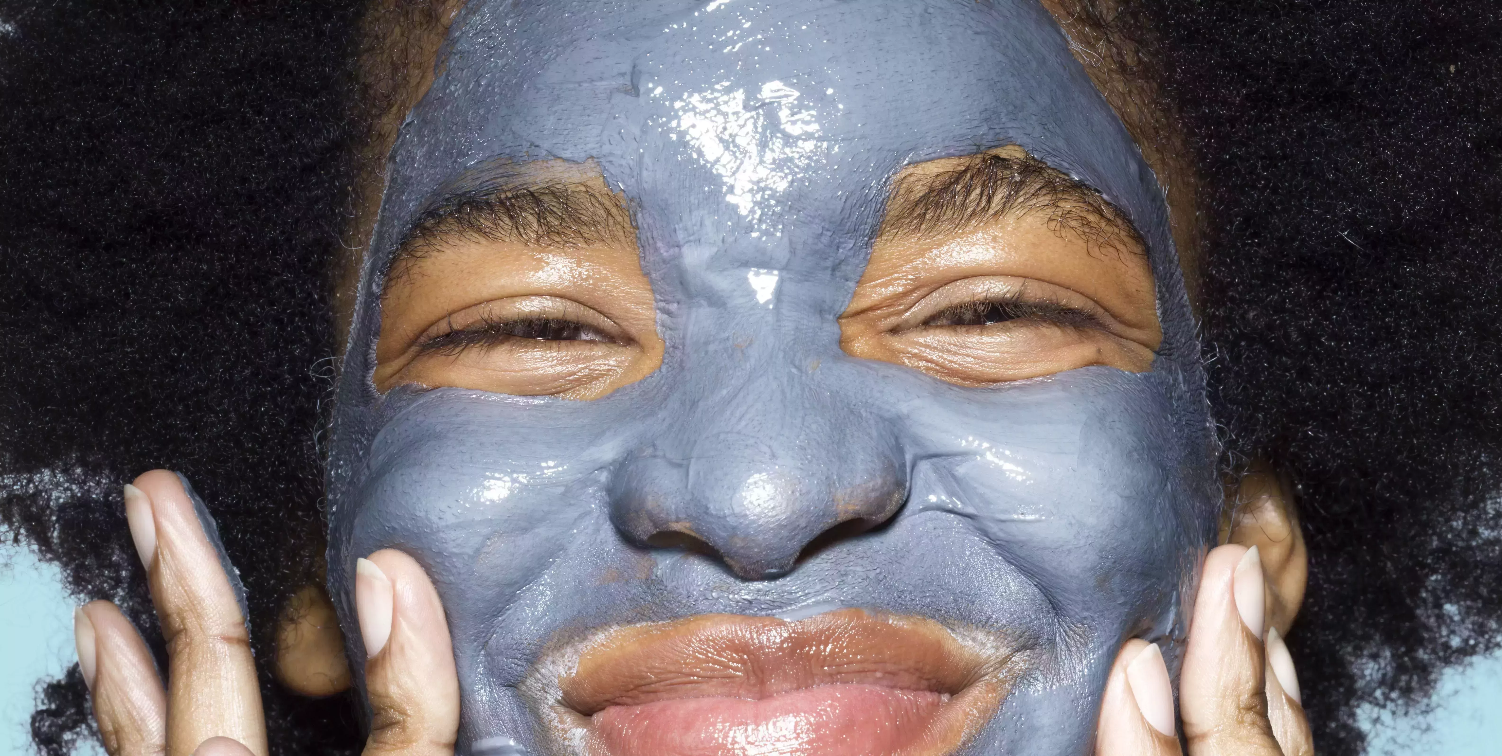 close up of young mixed race woman applying blue cleansing face mask on her face, with turquoise background